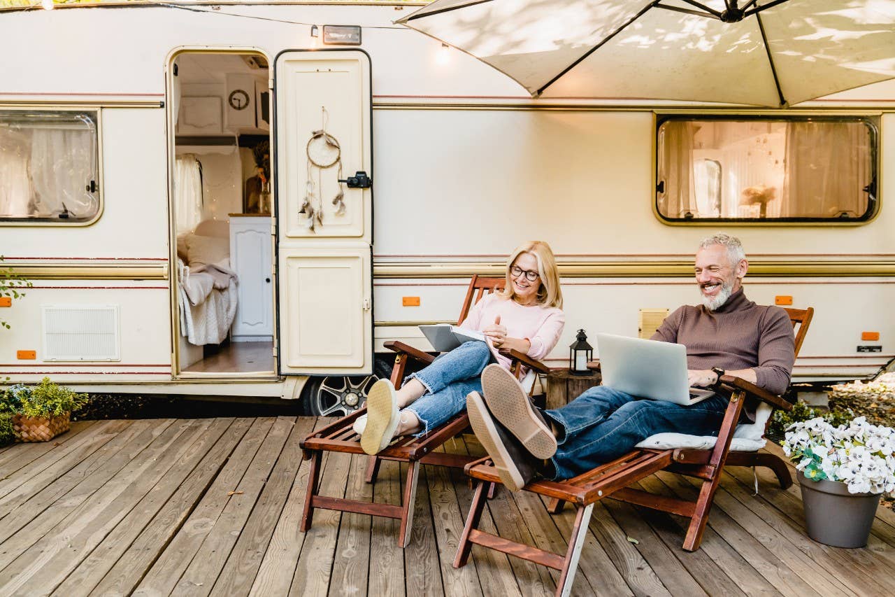 Pleased happy aged couple reading a book and using laptop while relaxing on the deck chairs near the camper van